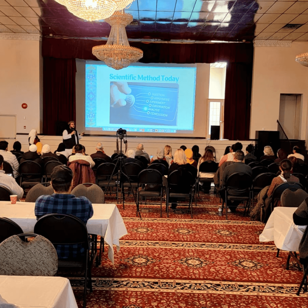 Audience seated in a hall watching a presentation on the scientific method projected on a screen.
