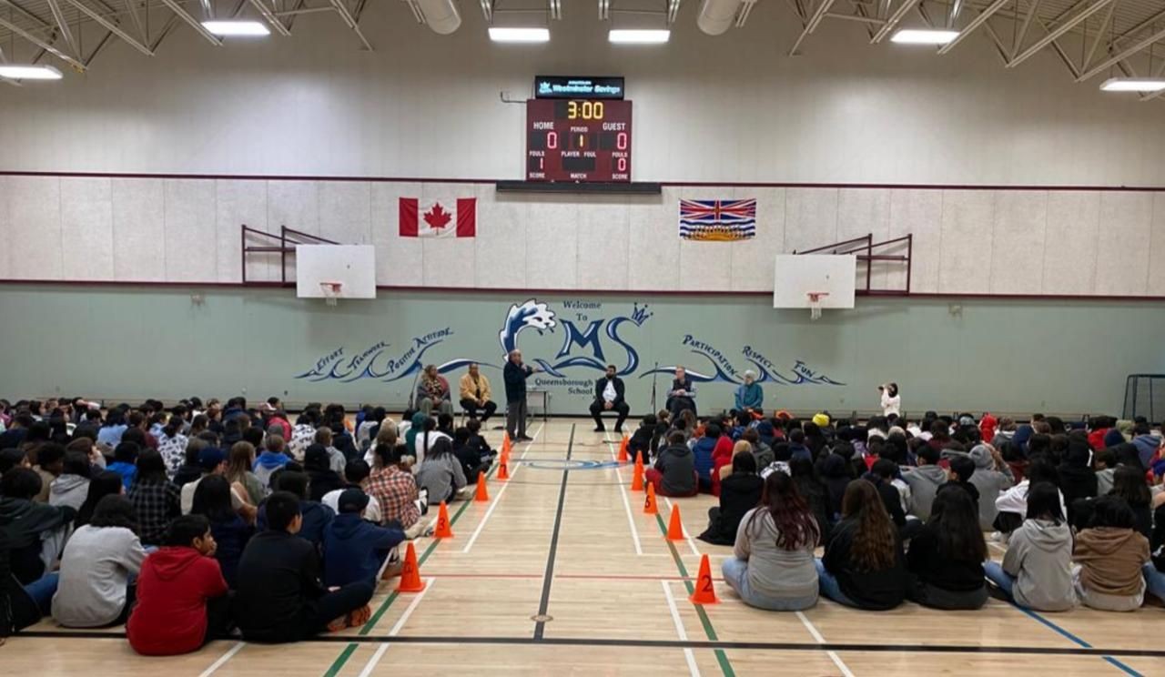 Students seated in a school gym with speakers at the front, Canadian and provincial flags displayed above.