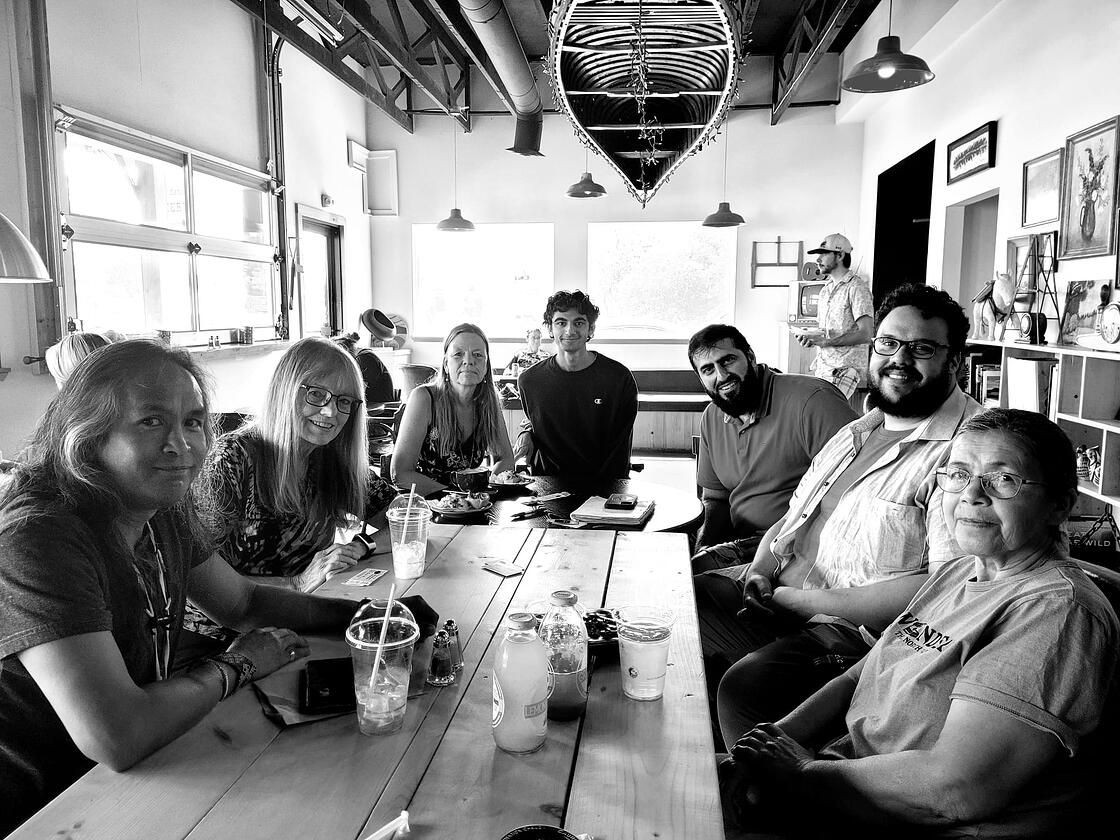 Group of people sitting at a long table in a cafe, talking and smiling.