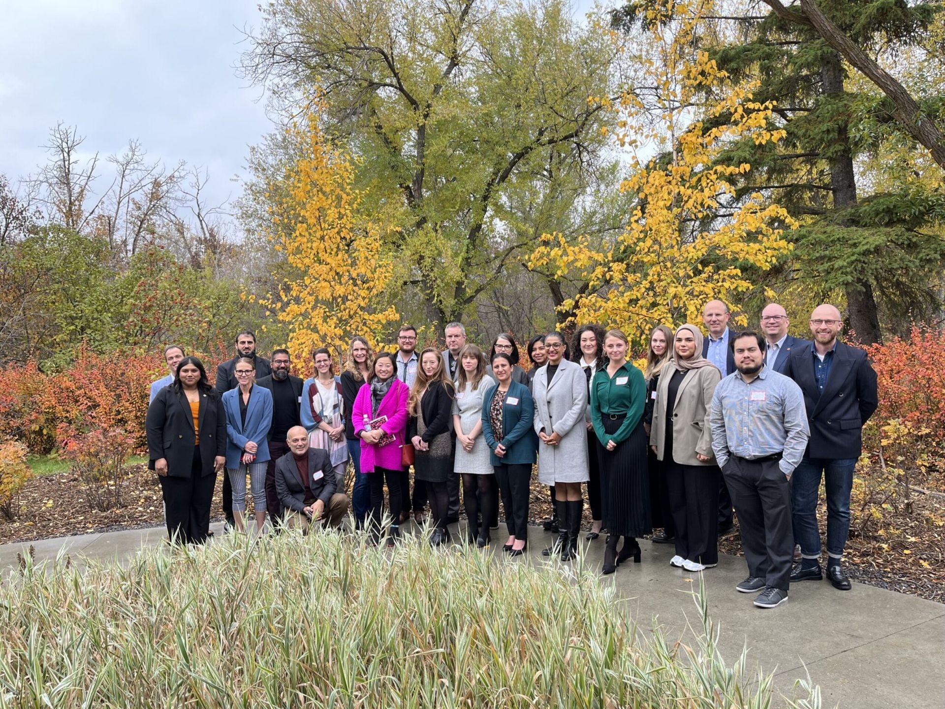 Group of people standing outdoors in a park with colorful autumn foliage and trees in the background.