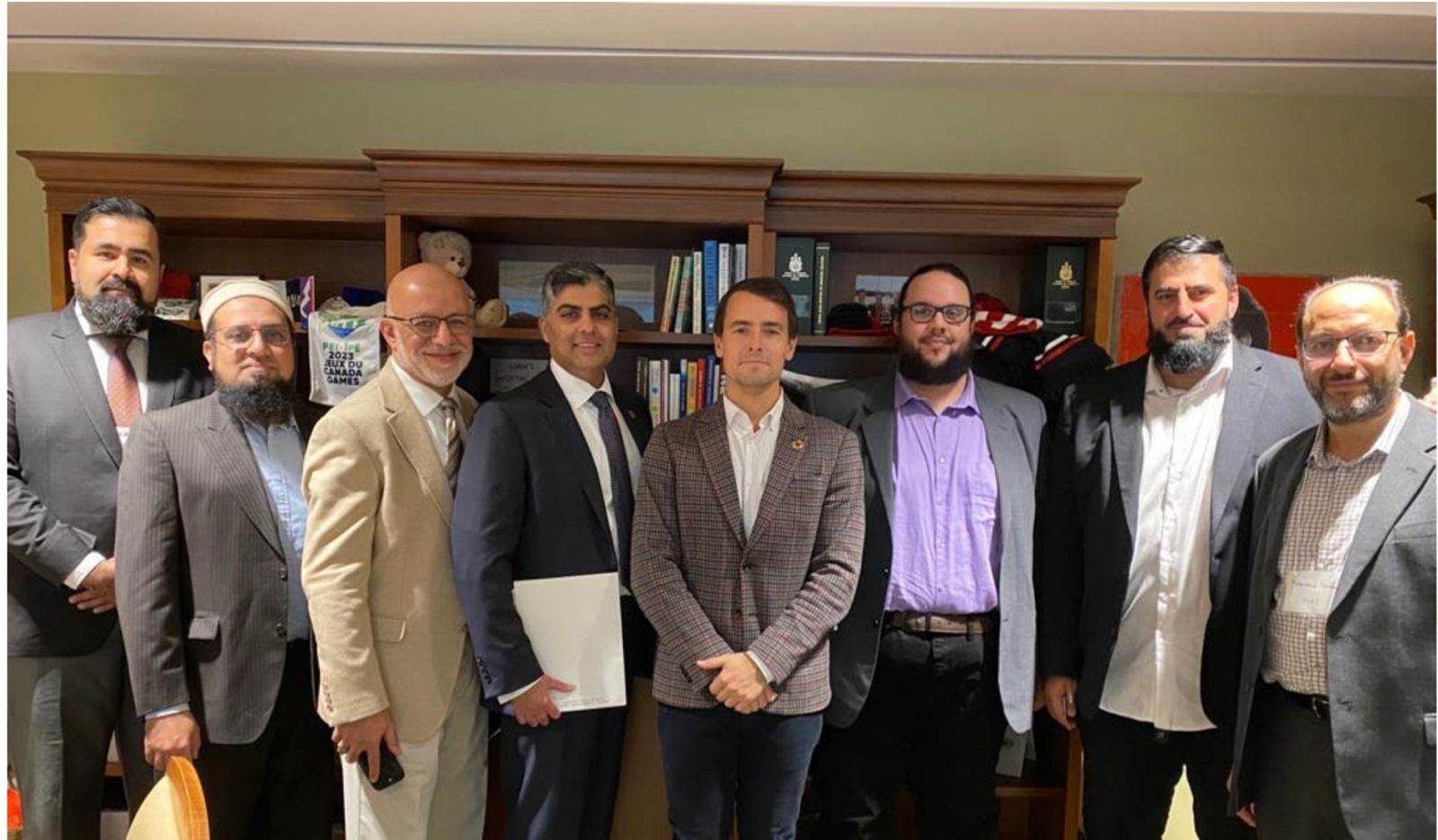 A group of eight men in suits standing in an office with bookshelves in the background.