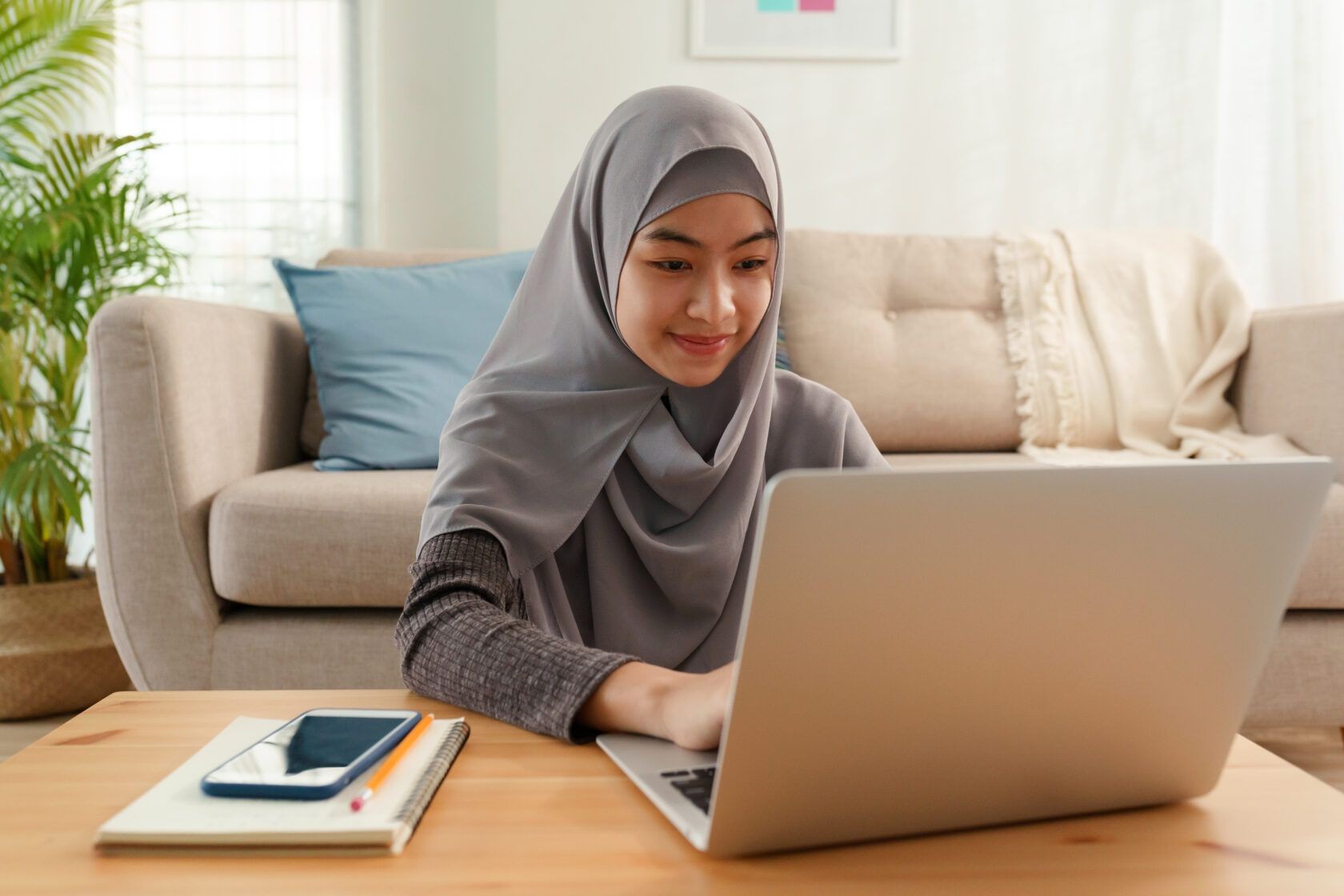 Woman wearing a hijab working on a laptop at home with a smartphone and notebook on table.