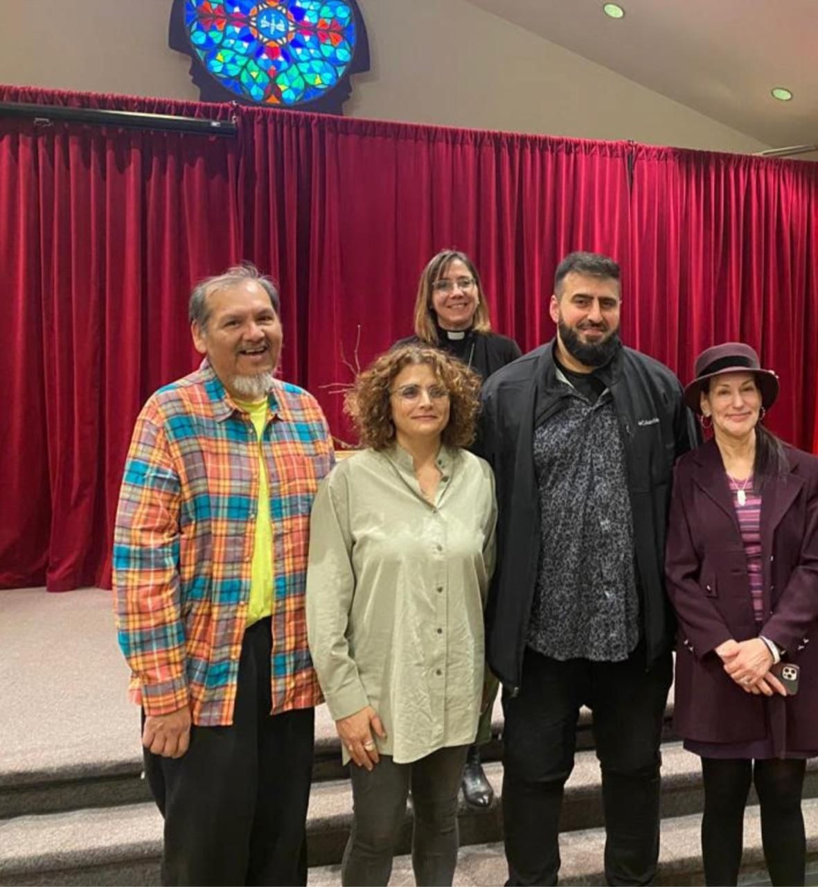 Group of five people standing together in front of red curtains and stained glass.