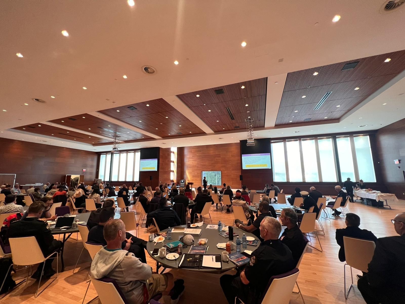 Large conference room with people seated at tables, facing a presentation screen.