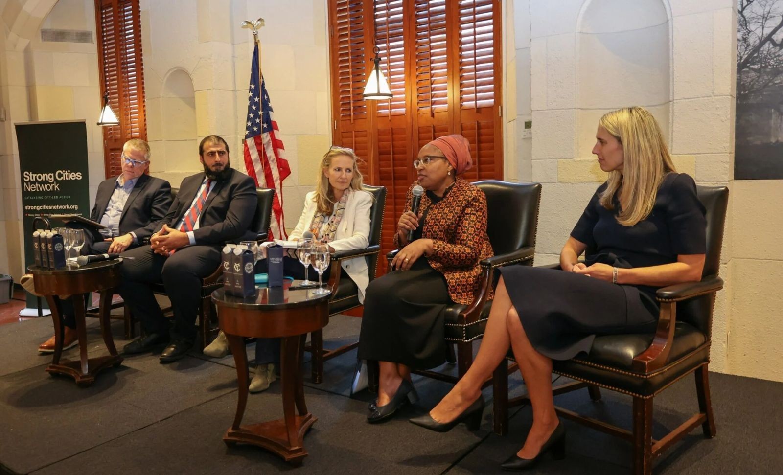 Panel discussion at Strong Cities Network event with five speakers seated on stage, American flag in background.