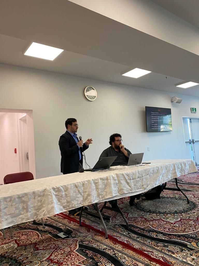 Two people at a conference table with laptops, one speaking into a microphone.